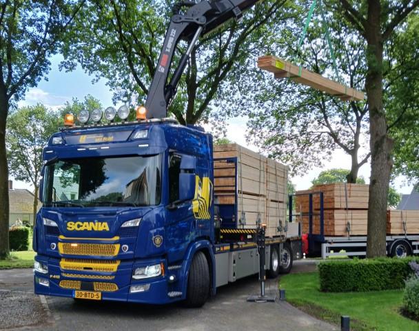 Een blauwe Scani met het bedrijfslogo lost een pakket hout bij een particulier in de tuin. 